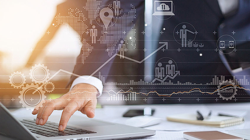 Das Bild besteht aus zwei Ebenen. Die vordere Ebene zeigt unterschiedliche Datenvisualisierungen: verschiedene Charts und Symbole. Die Ebene dahinter zeigt die Hand einer Person im Anzug und einen Laptop.