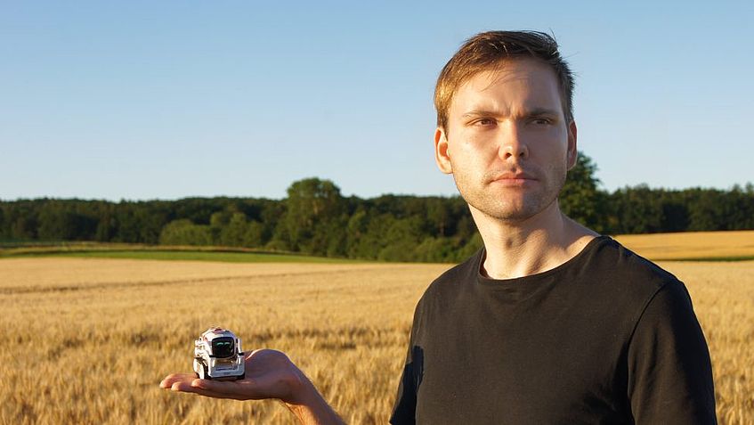 Mathias Weinhofer steht vor einem Getreidefeld und blickt an der Kamera vorbei in die Ferne. In der Hand hält er etwas, das wie ein kleiner Roboter oder eine Drohne aussieht.