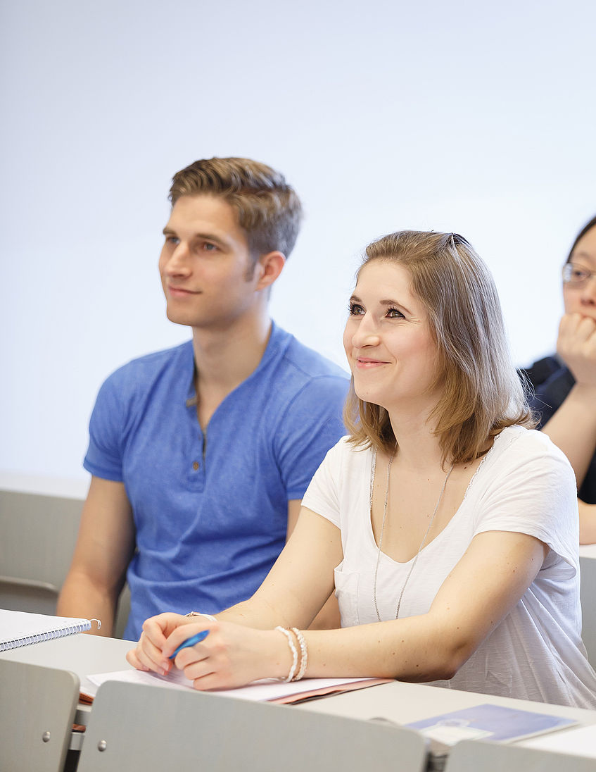 Zwei Studierende sitzen in einem Seminarraum und lächeln. Ihr Blick ist nicht direkt in die Kamera gerichtet, sondern vermutlich zu Vortragenden.