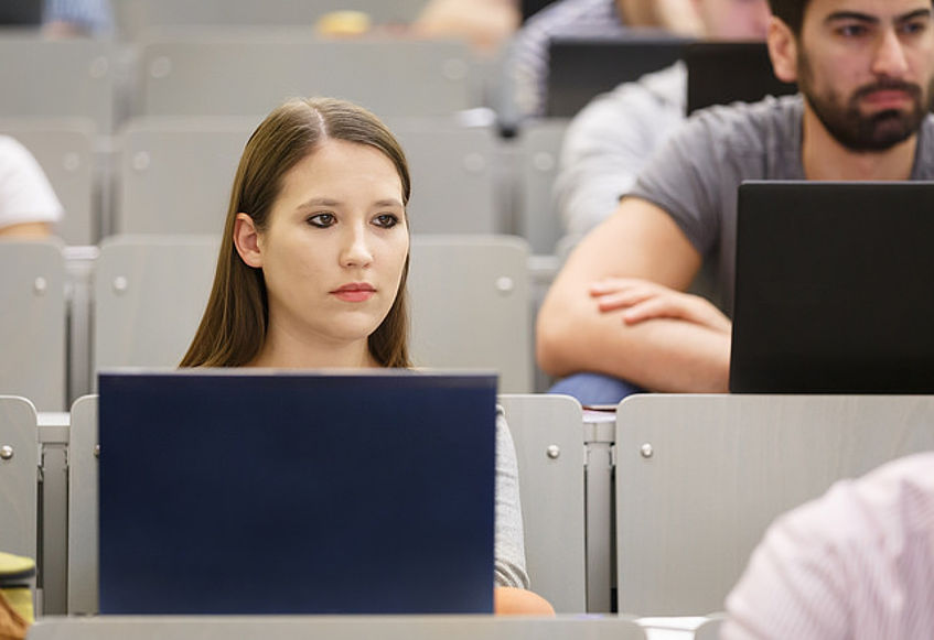 Eine Studentin sitzt im Hörsaal und schaut konzentriert nach vorne. Vor ihr steht ein Laptop. Im Hintergrund sind noch weitere Studierende sichtbar.